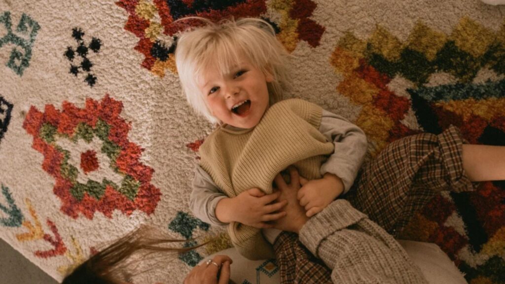 A high-angle, candid lifestyle shot of a laughing young boy with blonde hair lying on a vibrant patterned rug, captured with the Sigma 15mm F1.4 DC | Contemporary lens. This image demonstrates the lens's ability to capture sharp, emotional moments in low-light indoor settings while maintaining a wide-angle perspective. A versatile choice for family and documentary photography, this lens is available through Sigma Pakistan official and Hope Enterprises, the authorized distributor.