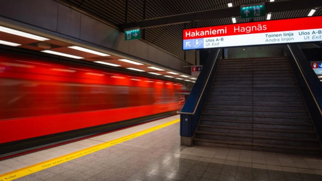 A long-exposure interior shot of the Hakaniemi metro station in Helsinki, featuring a blurred red train in motion against the sharp architectural lines of the stairs and ceiling. Captured with the Sigma 15mm F1.4 DC | Contemporary lens, this image demonstrates the lens's exceptional low-light performance and wide-angle perspective for urban and architectural photography. This high-performance prime lens is available through Sigma Pakistan official and Hope Enterprises, the authorized distributor.